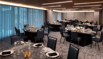 A modern banquet hall with round tables set for an event, featuring elegant tableware, flower centerpieces, and dim lighting from overhead fixtures.