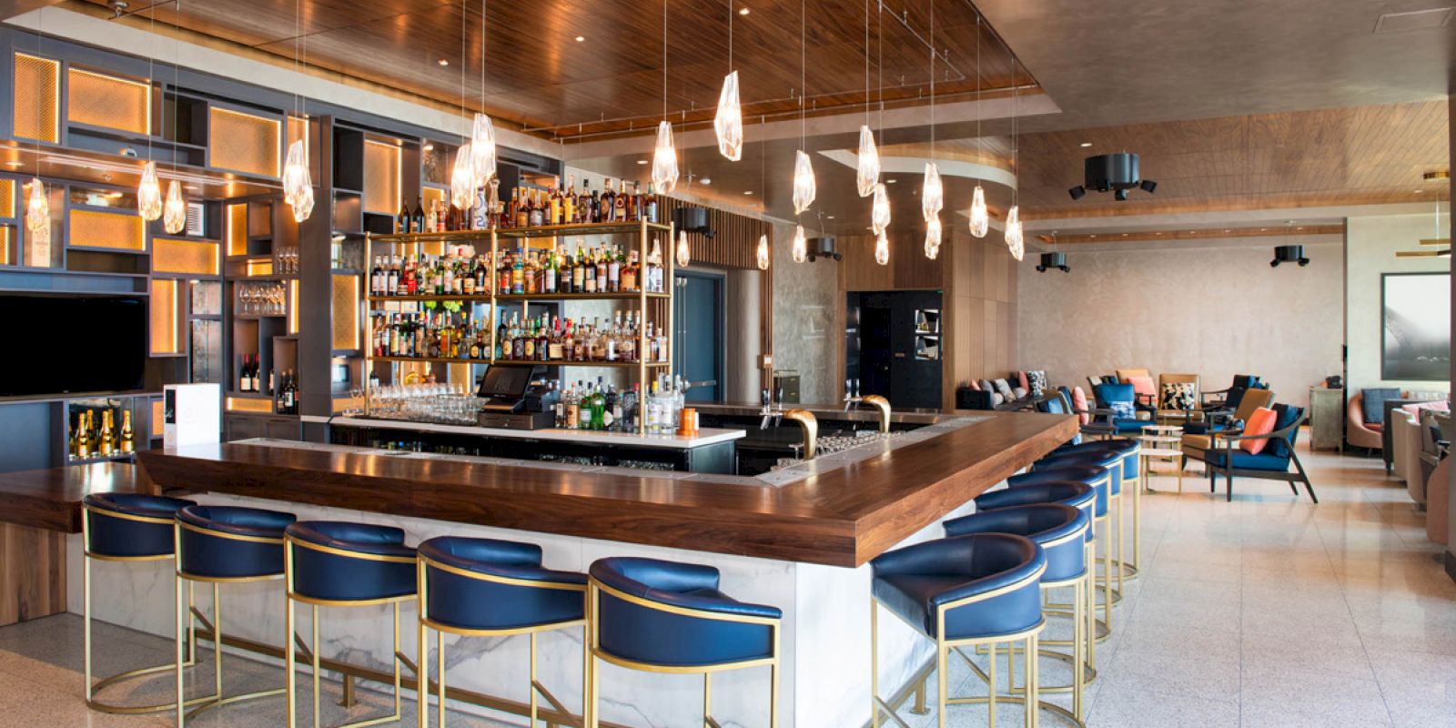 A modern bar with a wooden counter, blue barstools, and hanging pendant lights. The back shelves are stocked with various bottles. End of sentence.