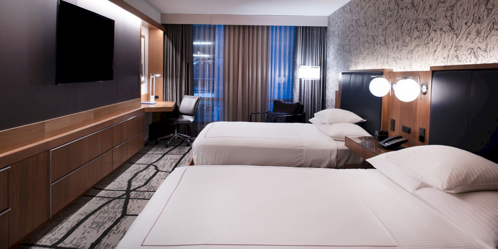 A modern hotel room with two neatly made beds, a wall-mounted TV, a desk by the window with a chair, and contemporary decor and lighting.