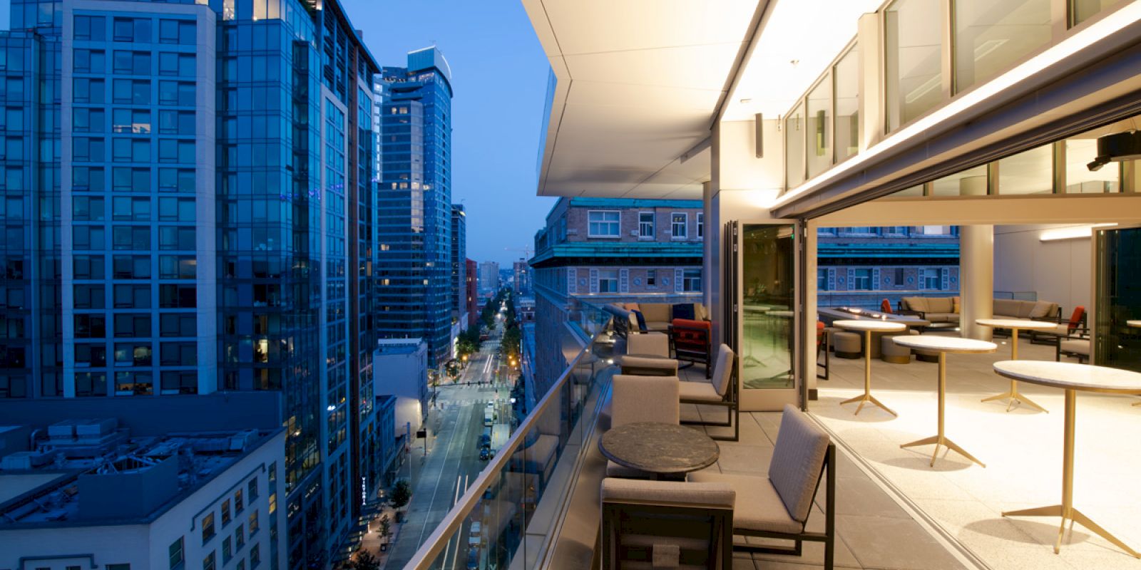 A modern balcony overlooking a city street at dusk, with outdoor seating and tables. High-rise buildings are visible in the background.