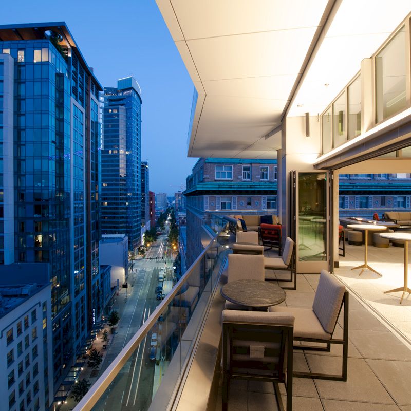 A modern cityscape at dusk with tall buildings, a brightly lit outdoor patio featuring tables and chairs, and an open balcony with glass railings.