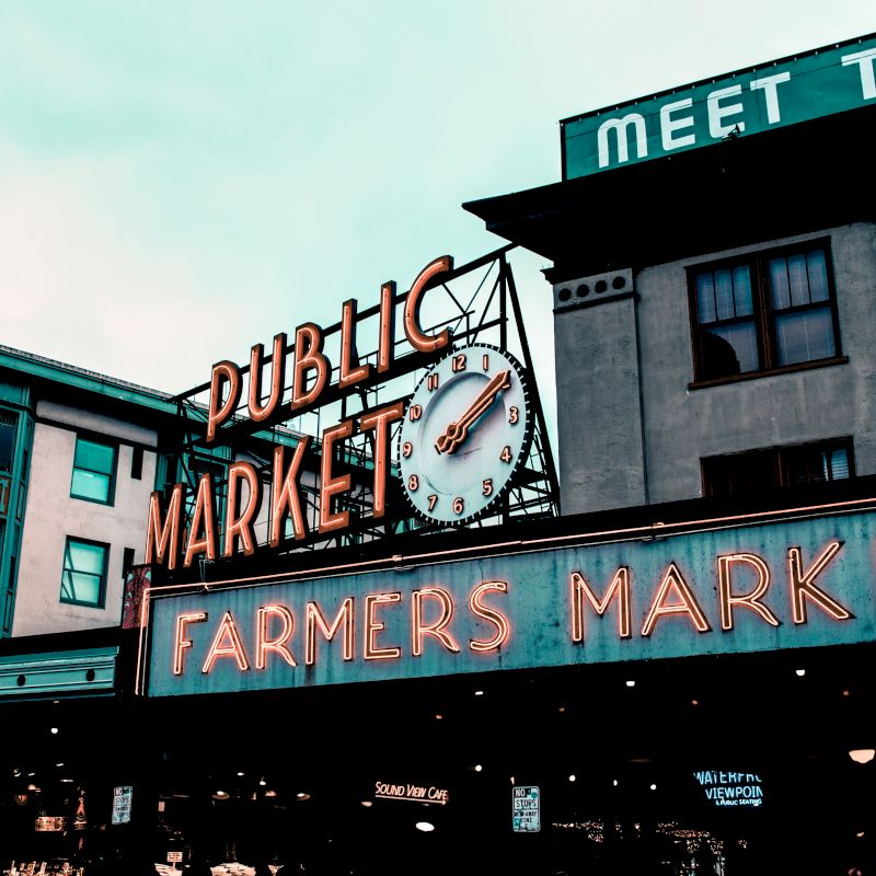 This image features a neon sign for a "PUBLIC MARKET" and "FARMERS MARKET" with a clock beneath. Surrounding buildings have visible signage.