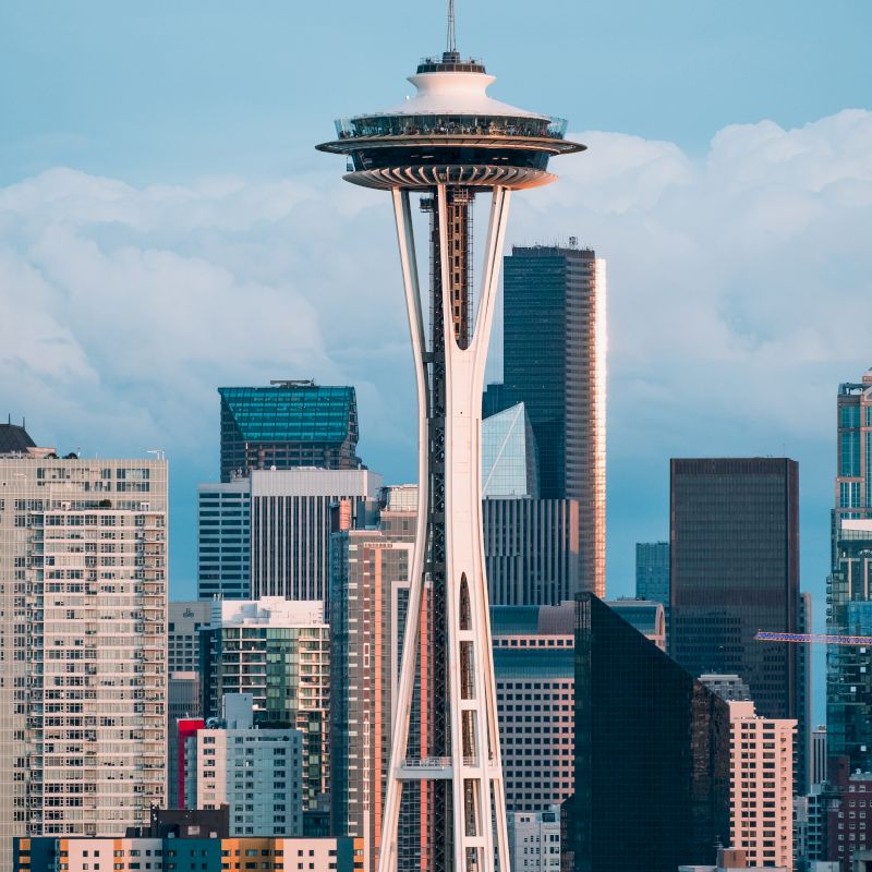 The image shows the Space Needle in Seattle, with the cityscape and various skyscrapers in the background under a blue sky with clouds.