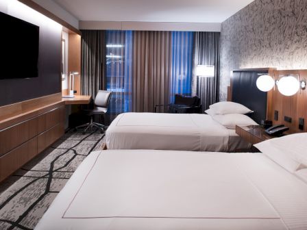 The image shows a modern hotel room with two beds, a TV on the wall, a desk, a chair, and stylish decor with patterned wallpaper and curtains.