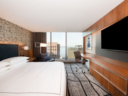 A hotel room with a large bed, a wall-mounted TV, a work desk and chair, and a window offering a city and water view.