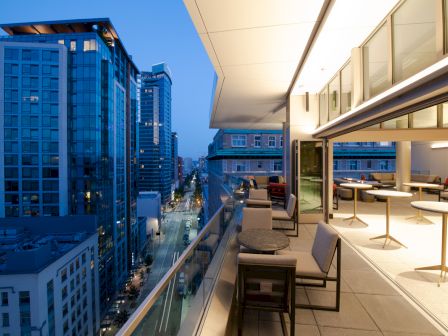 A modern cityscape at dusk is visible from an outdoor terrace with tables and chairs, framed by glass buildings and a vibrant city street below.