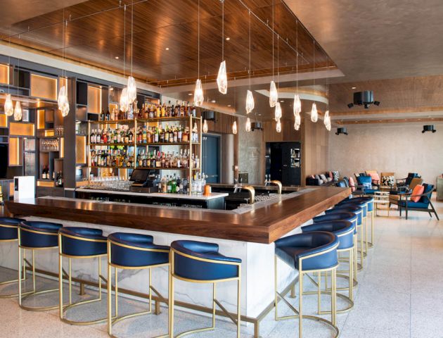A modern bar with stylish seating, pendant lights, and a well-stocked liquor display is visible. The seating area has a cozy, contemporary design.