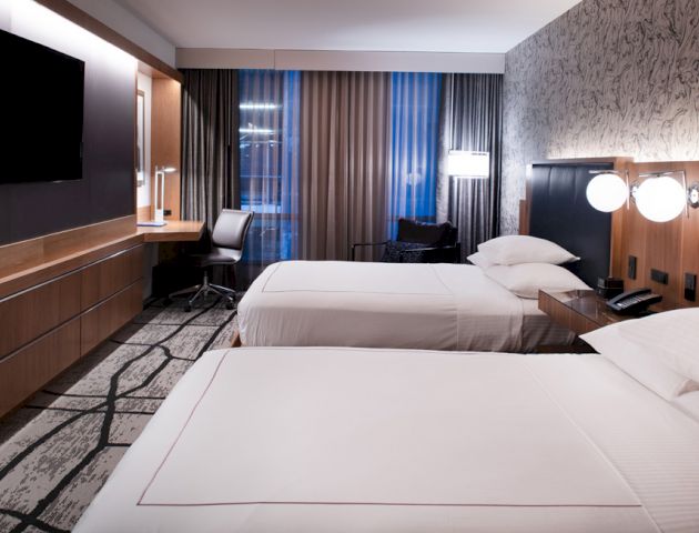A modern hotel room with two neatly made beds, a wall-mounted TV, a desk with a chair, and patterned wallpaper.