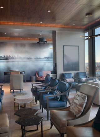 A modern lounge area with cushioned chairs, tables, a cityscape mural, and large windows offering a view of the waterfront and Ferris wheel.