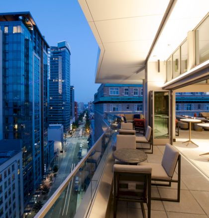 The image shows a modern city view from a stylish balcony with tables and seating, overlooking high-rise buildings and a street below, during dusk.
