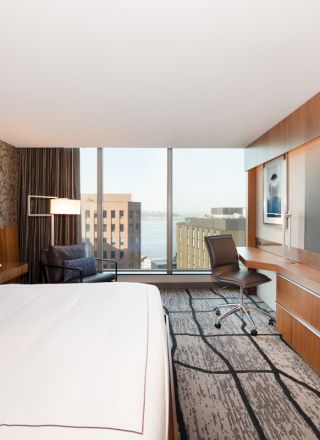 A modern hotel room with a large bed, a flat-screen TV, a desk, and a view of buildings and water through the window.