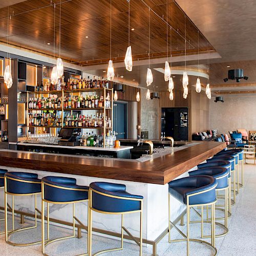 A modern bar with a wooden countertop, blue cushioned stools, pendant lights, and shelves filled with bottles, in a spacious lounge area.