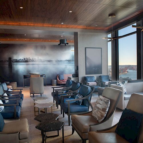 A modern lounge area with large windows overlooking a waterfront, numerous chairs, and a wall mural depicting a city skyline at sunset.