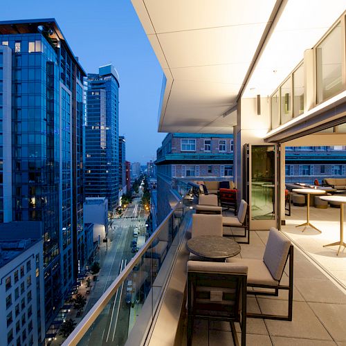An urban balcony view at dusk with city buildings, an empty outdoor seating area, and a lit interior space.