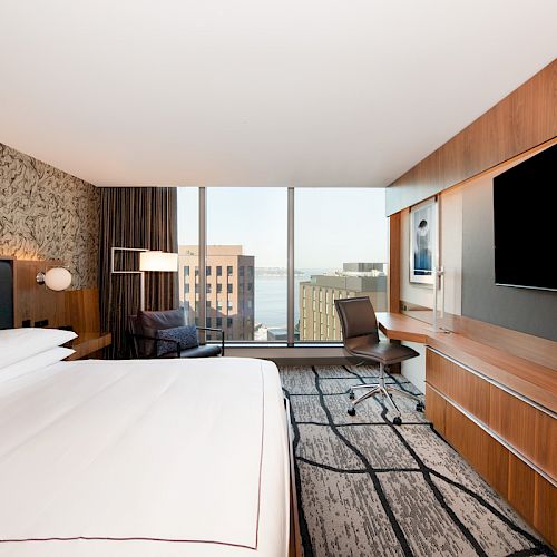 A modern hotel room with a large bed, desk, chair, TV, and window overlooking city buildings and a body of water. The room has stylish decor.
