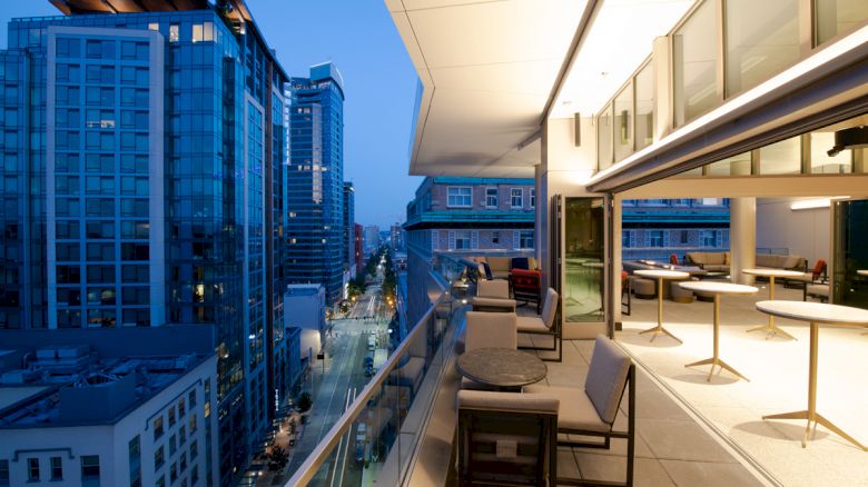 A modern urban balcony overlooks a city street with tall buildings. The balcony is furnished with chairs and tables, and it's evening time.