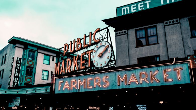This image shows the exterior of a public market with neon signs reading "PUBLIC MARKET" and "FARMERS MARKET." A clock is also visible.