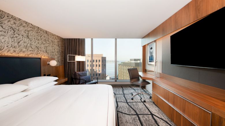 Modern hotel room with a large bed, stylish decor, flat-screen TV, desk, chair, and a city view through a floor-to-ceiling window.