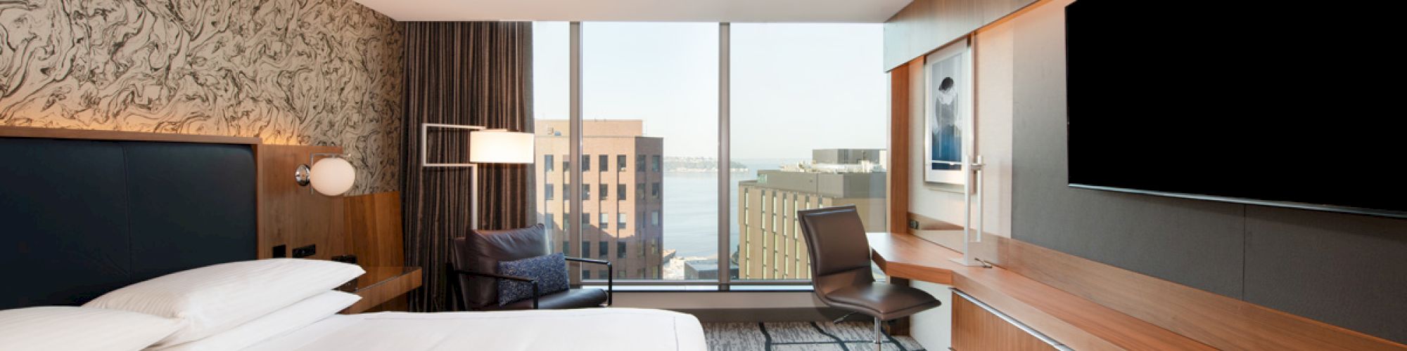 The image shows a modern hotel room with a large bed, a flat-screen TV on a wooden wall unit, a work desk and chair, and a view of buildings.