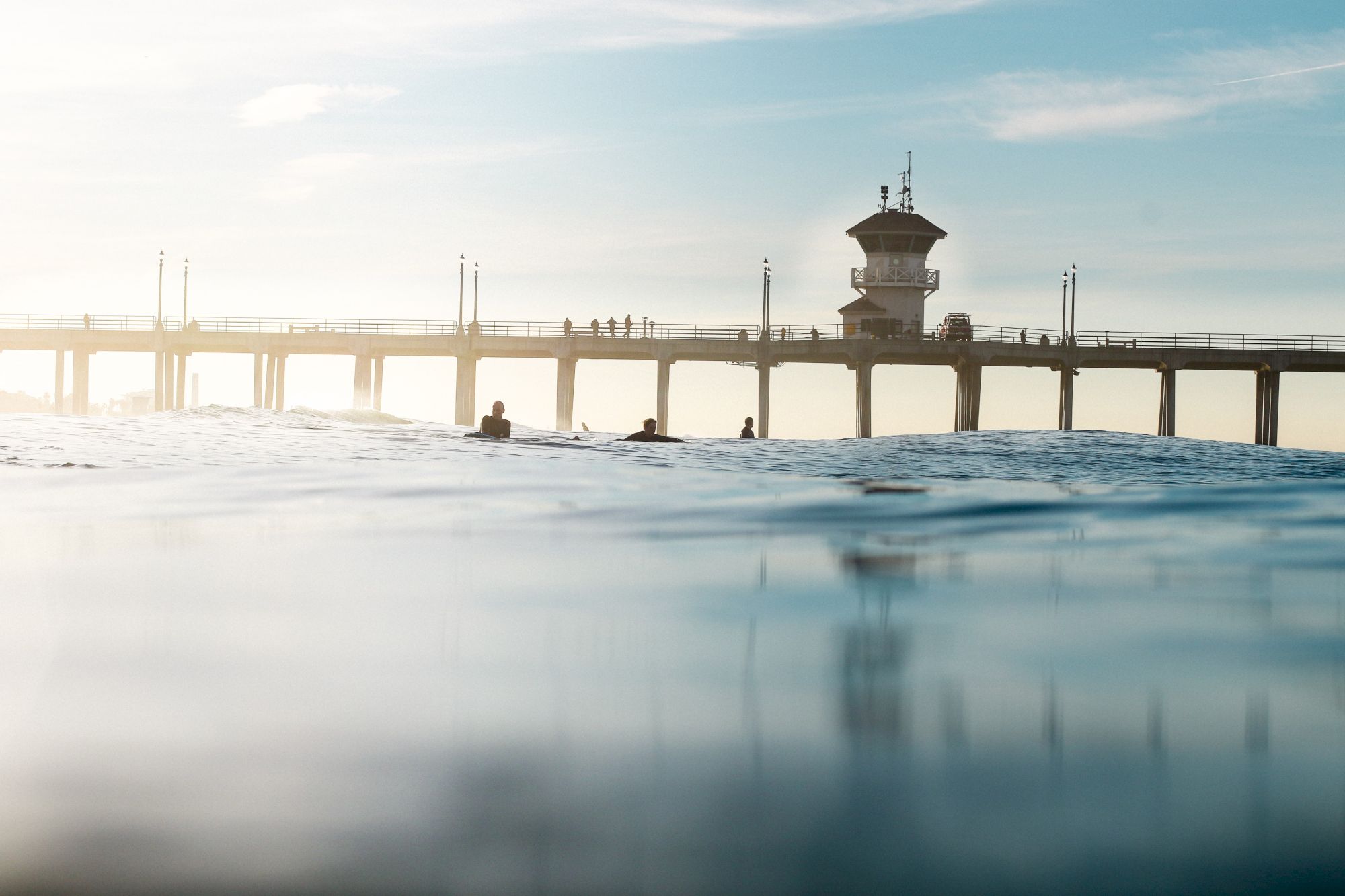 The image shows a coastal pier with people walking on it. The foreground features water and distant silhouettes of swimmers or surfers in the ocean.